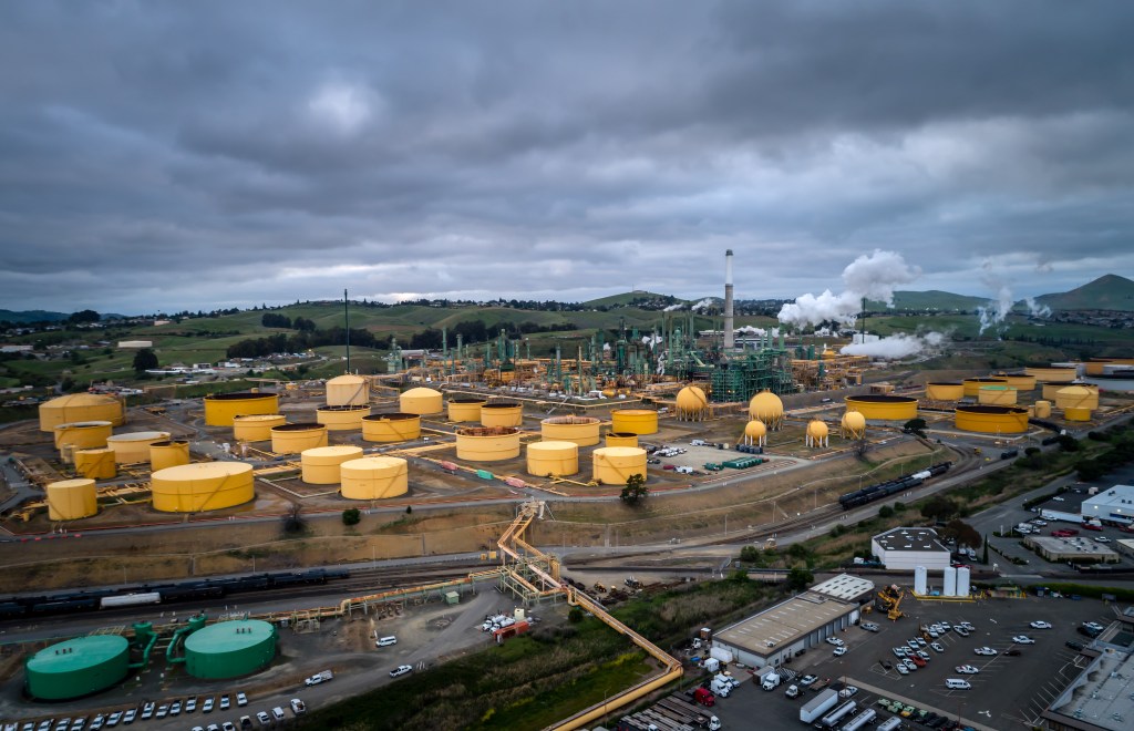 Aerial view of the Valero Refinery in Benicia, California, featuring large yellow and green storage tanks, industrial structures, and white smoke rising from chimneys, set against a backdrop of green hills and an overcast sky.