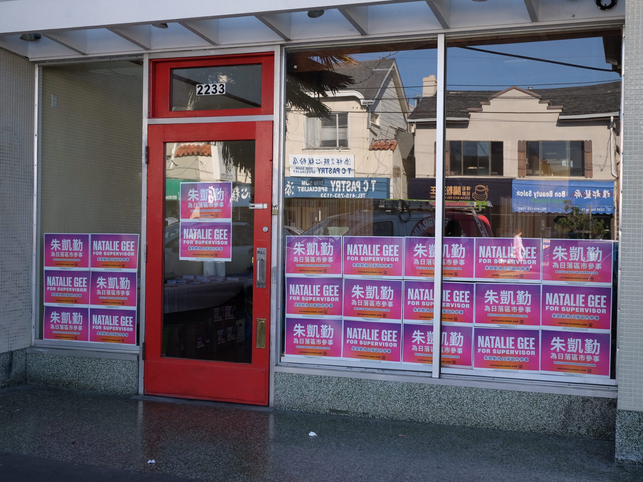 Storefront with a red door and large windows covered in pink campaign posters for Natalie Gee, candidate for supervisor, with both English and Chinese text.