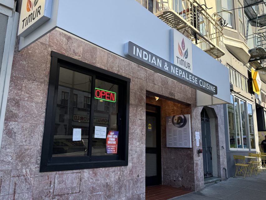 The exterior of a restaurant named Timur, featuring a sign for Indian & Nepalese cuisine, a neon "OPEN" sign, menu display, and a stone facade.