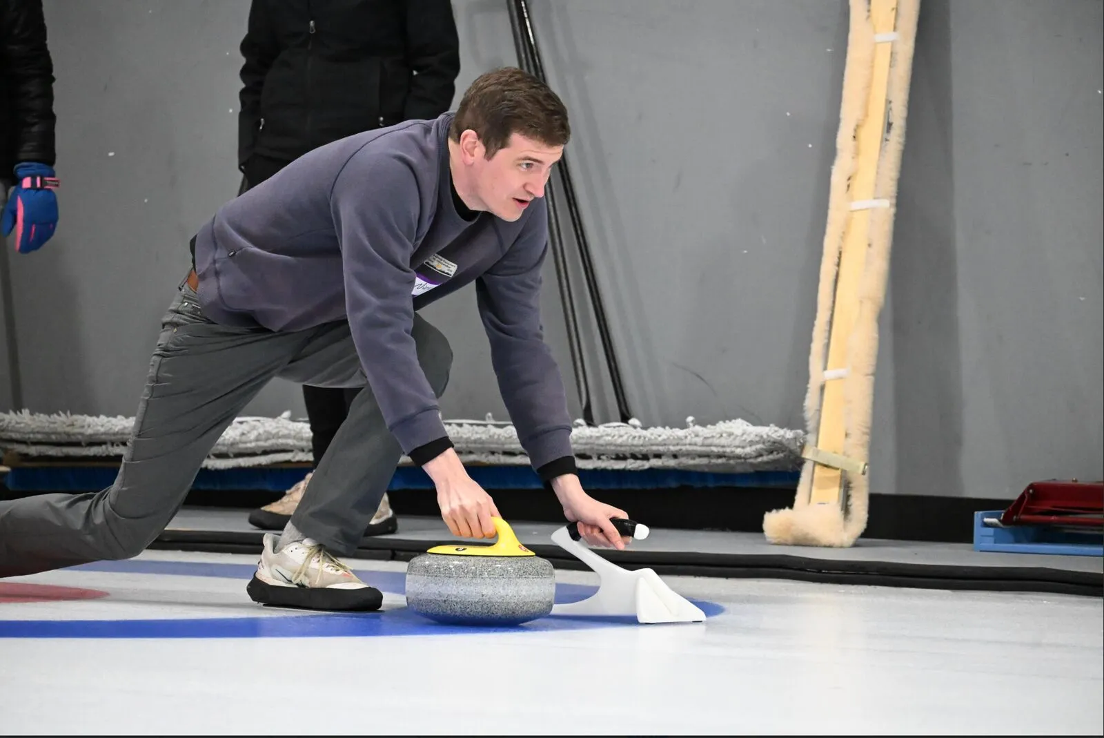 A man slides across ice in a crouched position pushing a curling stone with a focused look on his face.