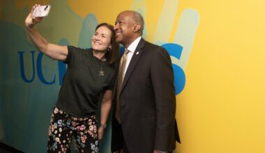 A woman and a man smiling together while taking a selfie against a colorful background.