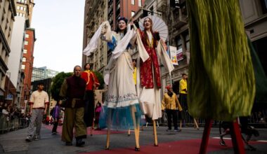 Two people dressed in decorative outfits walk on stilts in the street.