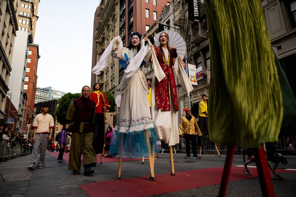 Two people dressed in decorative outfits walk on stilts in the street.