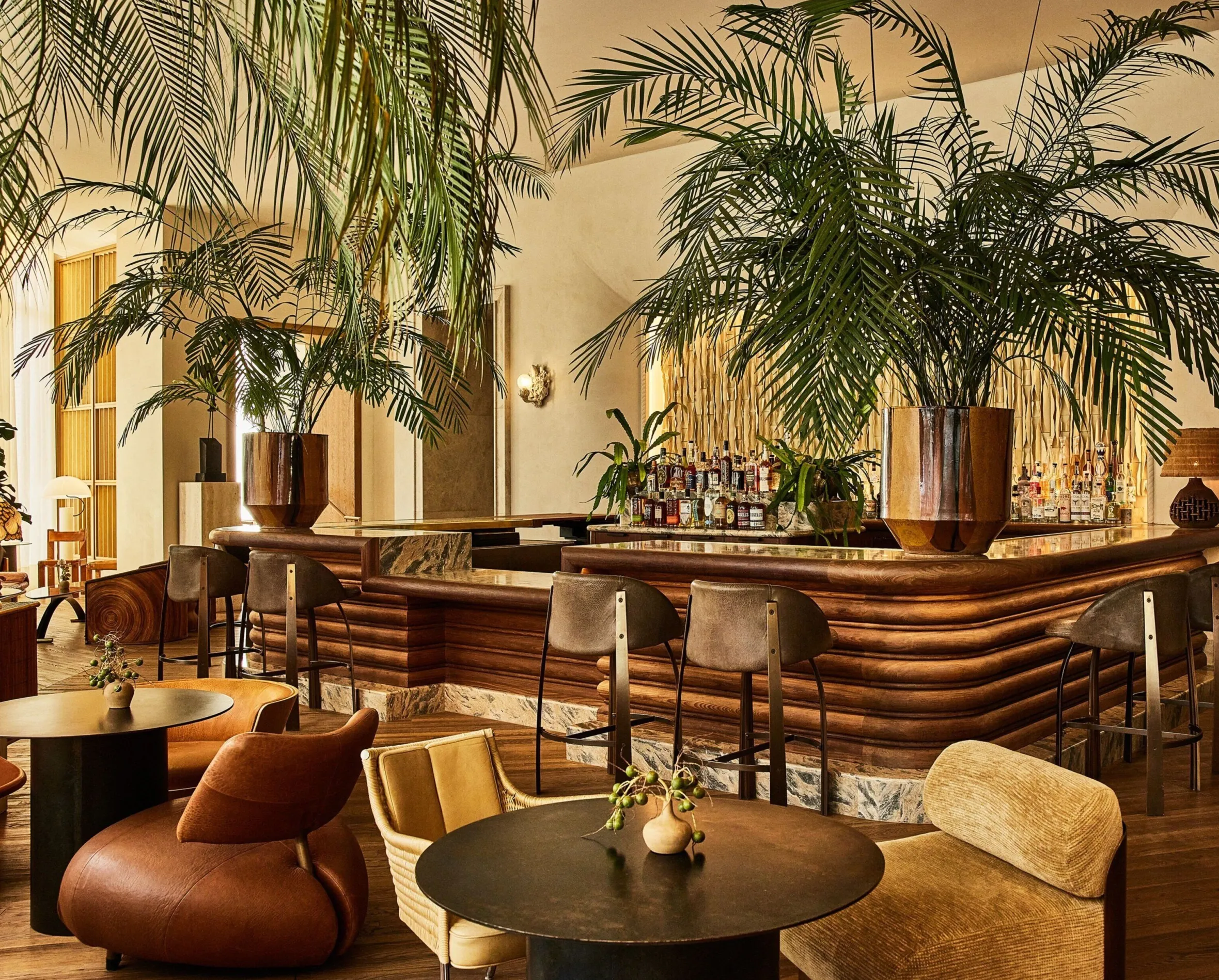 Interior view of the Santa Monica Proper Hotel bar, with a wooden bar, bar stools, and seating arrangements, complemented by large palm plants.