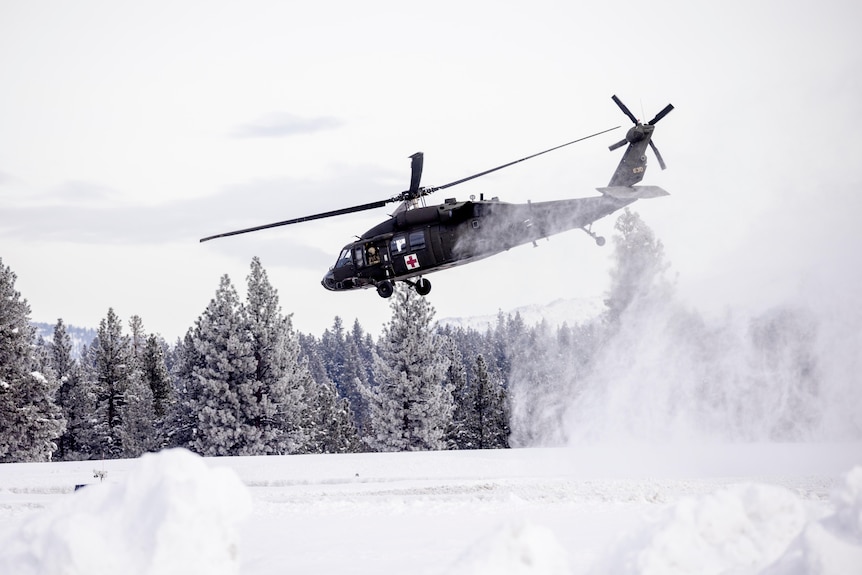 A black helicopter with a red cross takes off from a snowy field.