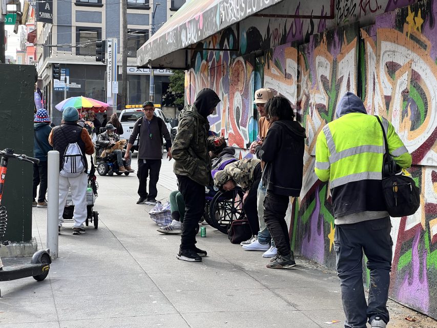 A group of people gather near a graffiti-covered wall on 16th St. Plaza. Some chat while others sit on the ground, soaking in the urban vibe. Pedestrians and a person with a walker weave through the bustling city scene in the background.