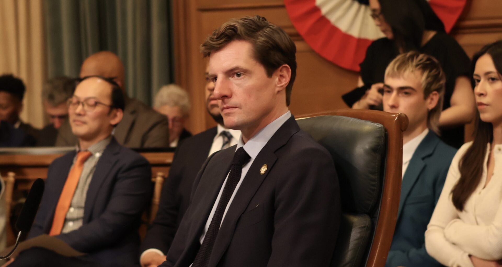 A group of people in business attire sit attentively in a government meeting room, with a large, patriotic banner in the background.