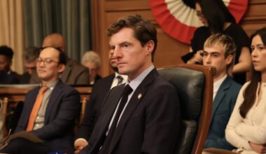 A group of people in business attire sit attentively in a government meeting room, with a large, patriotic banner in the background.