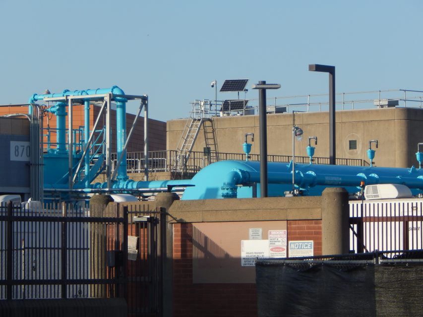 Industrial facility with large blue pipes, fencing, warning signs, and various structures under a clear blue sky.