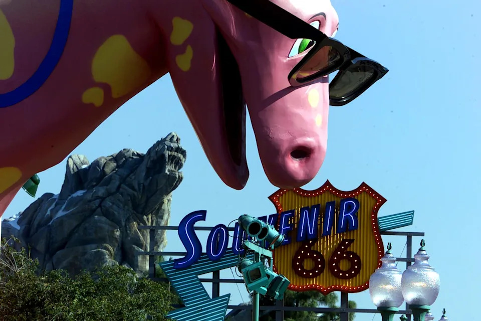A pink dinosaur in sunglasses in a theme park, with a Route 66-themed shop in the background.
