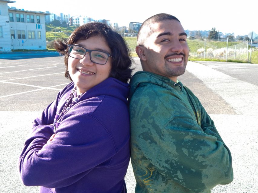Two people stand back-to-back outdoors, both smiling at the camera. One wears a purple hoodie and glasses, the other wears a green hoodie.