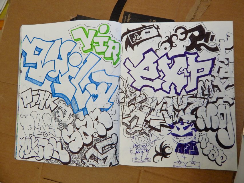 Open sketchbook showing colorful and black-and-white graffiti-style drawings, including bold text, cartoon faces, and abstract designs, placed on a cardboard surface.