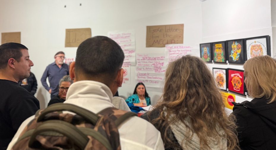 A group of people are gathered in a room with handwritten signs and artworks on the walls; a woman is seated at a table, appearing to address the group.