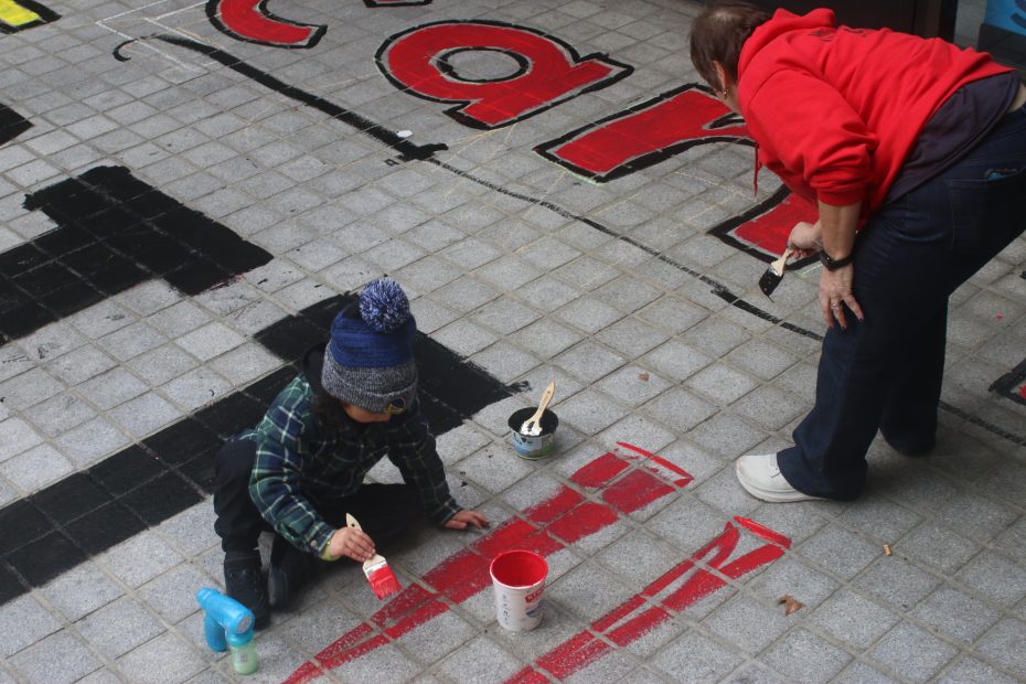 A child and an adult paint large red and black letters on outdoor gray tiles. The child sits and paints with a brush, while the adult stands, also holding a brush.