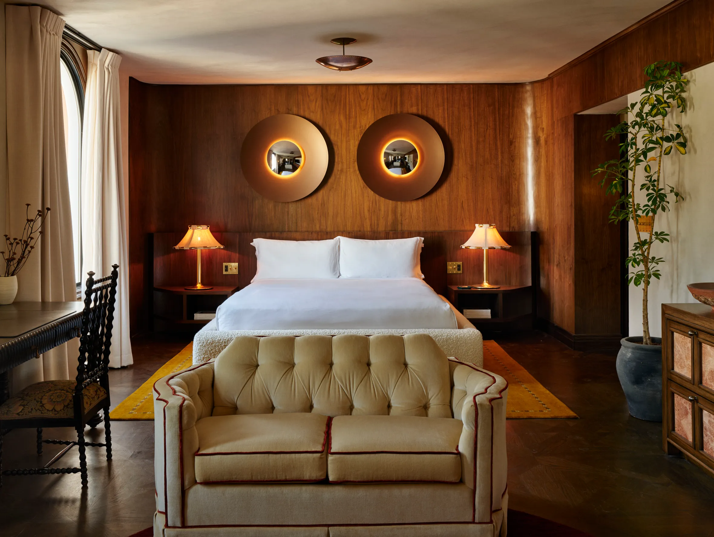 A bedroom at The Hollywood Roosevelt with a bed, desk, couch, and plant, featuring wooden walls and warm lighting.