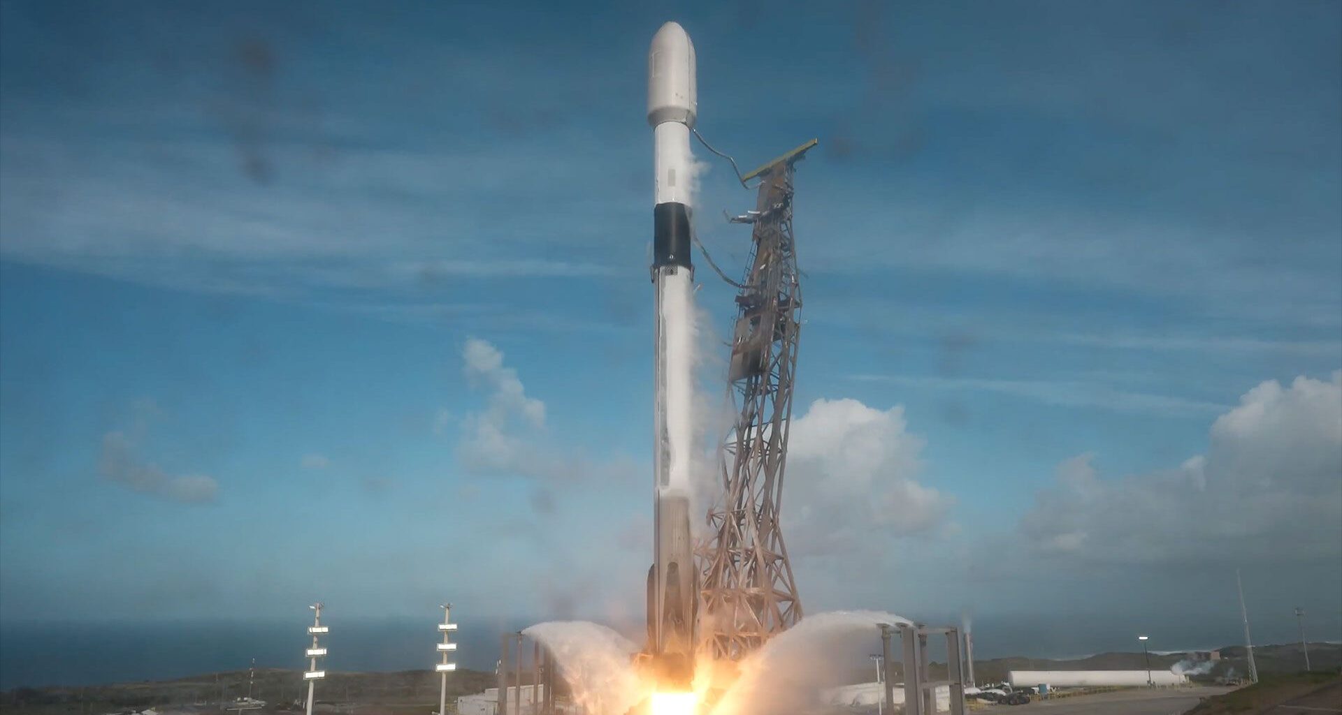 A white and black rocket lifts off from its launch pad into a partially cloudy blue sky.