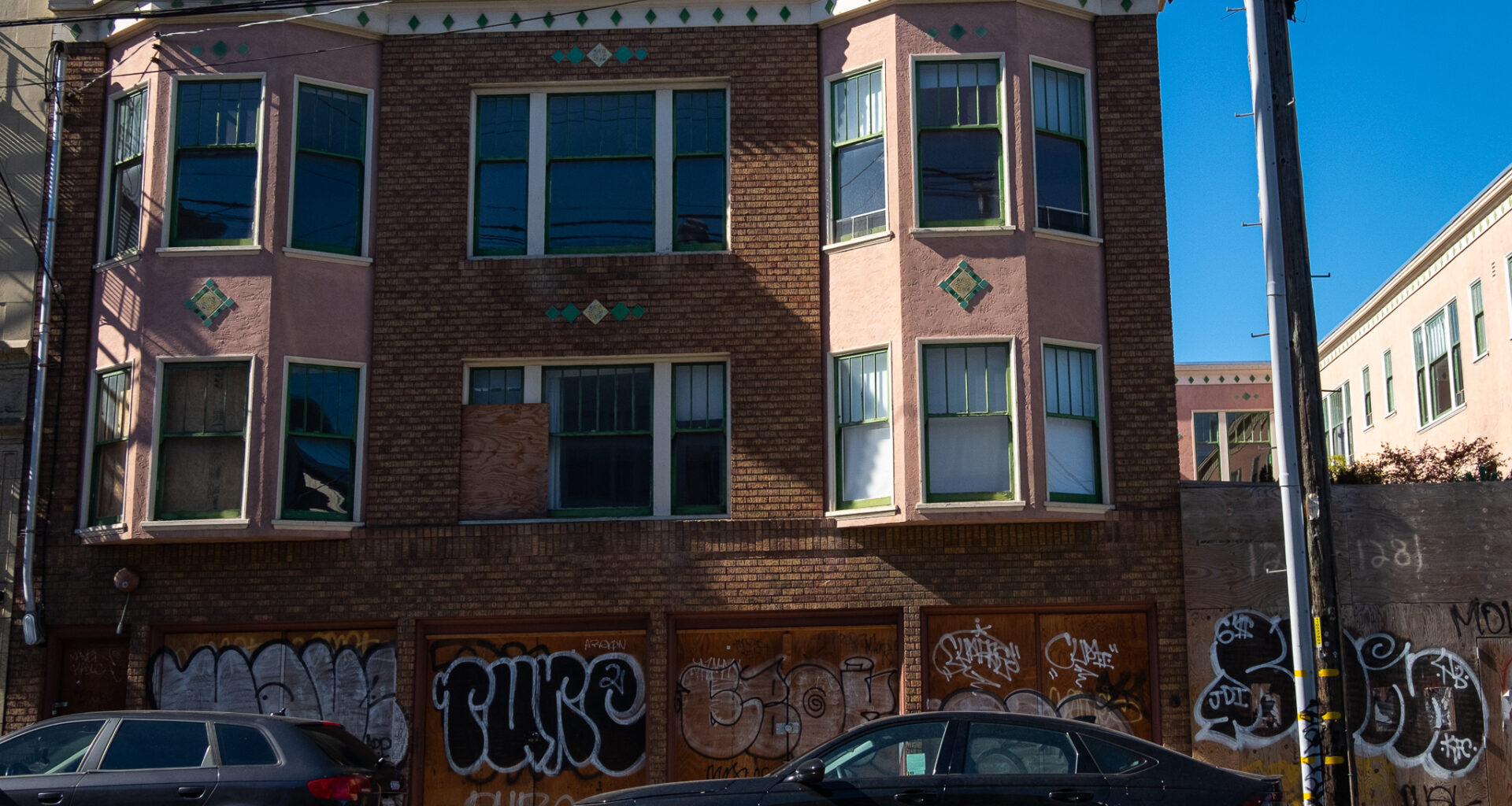 A two-story brick and stucco apartment building with boarded-up windows and graffiti on the ground floor; two parked cars are in front.