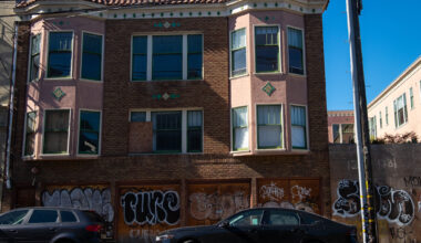 A two-story brick and stucco apartment building with boarded-up windows and graffiti on the ground floor; two parked cars are in front.