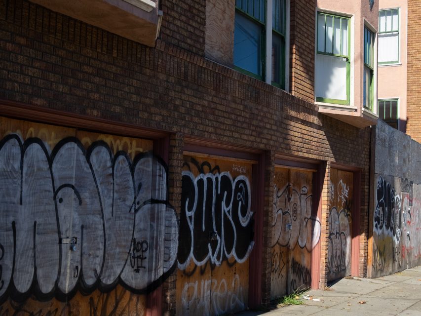 Row of brick building garage doors covered in graffiti on a sunny day; some windows above have partially open blinds.