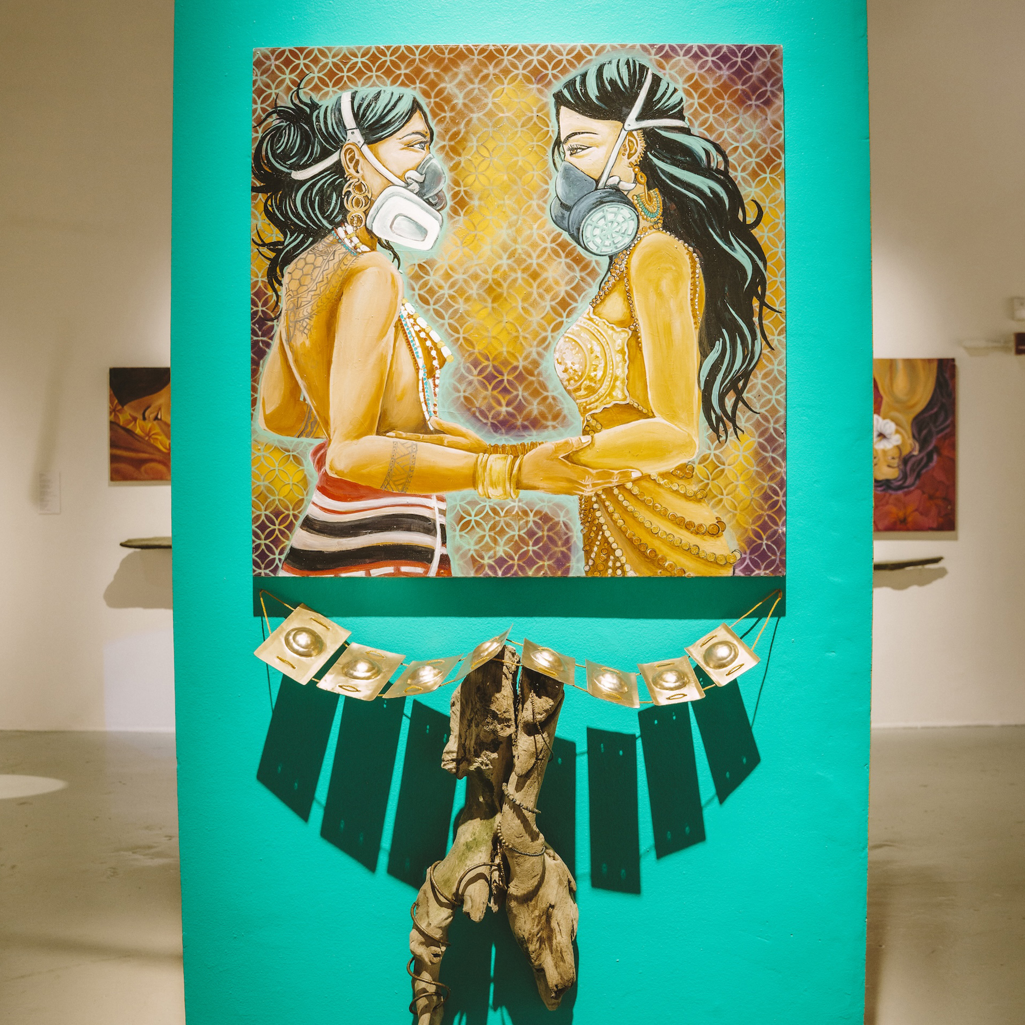 A painting of two women wearing masks, facing each other, mounted on a green background in an art gallery.