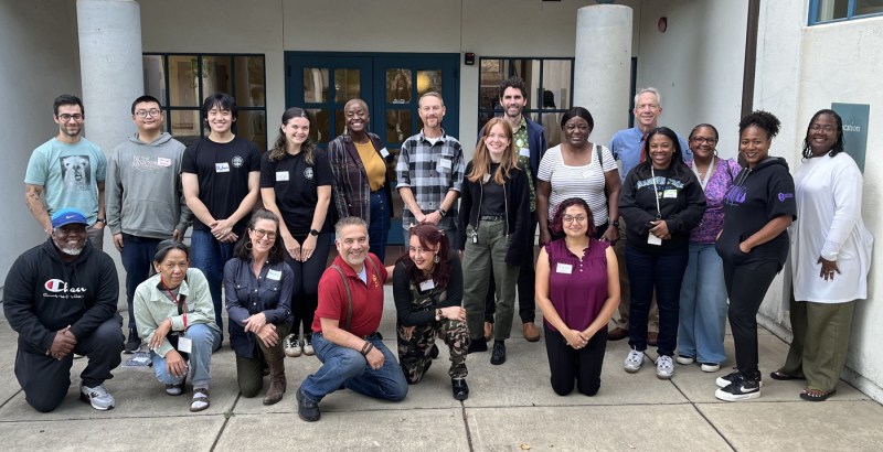 A diverse group of approximately 17 people pose together in two rows, some standing and some kneeling, in an indoor space with white columns and glass doors in the background.