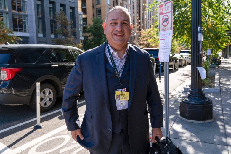 Smiling man in a suit stands on a sunny city street, wearing a badge and holding a bag. Urban buildings and parked cars are visible in the background.