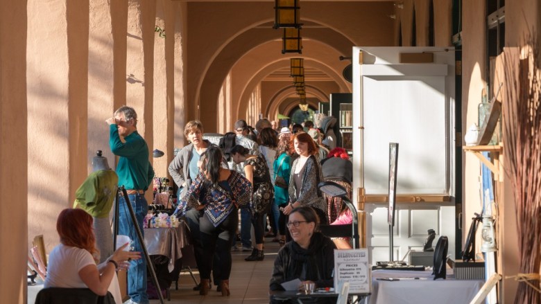 People at an outdoor market in an arched corridor with sunlight streaming through.