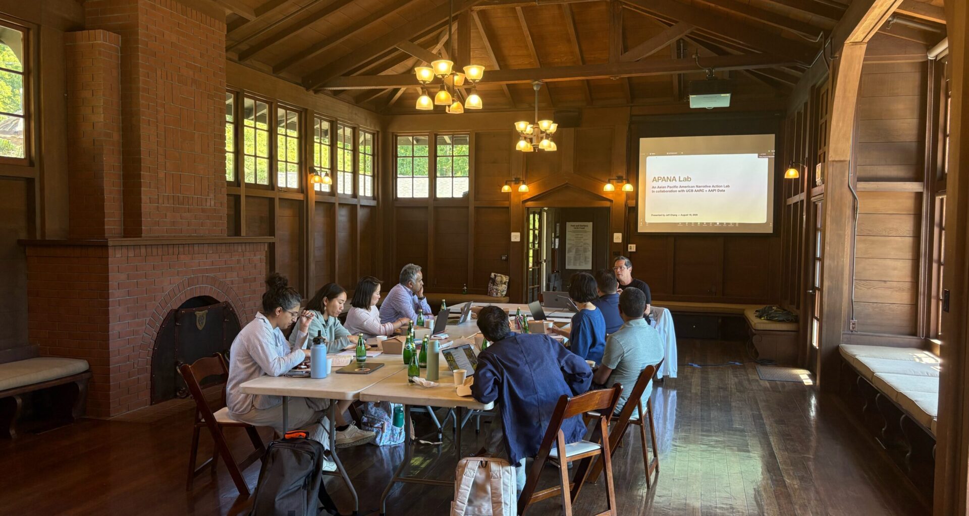 A group of faculty sit around a table in a wood-paneled room, looking at a screen