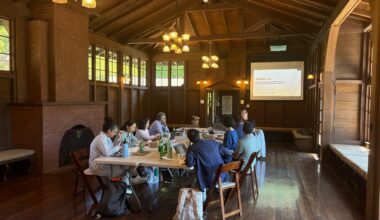 A group of faculty sit around a table in a wood-paneled room, looking at a screen