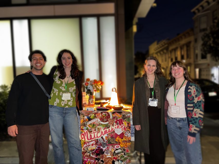 Four people stand and smile by a decorated table outdoors at night, celebrating Valentine's Day, with buildings and a lit lamp visible in the background.