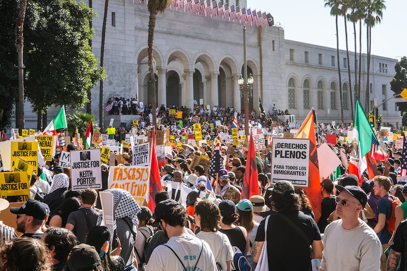 The street in front of the City Hall building is packed with protesters with signs and flags in the sun.