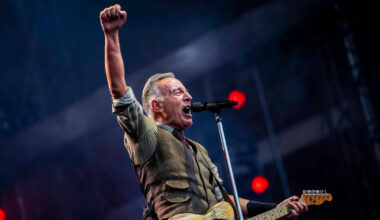 A man in a collared shirt and vest holds a Fender telecaster guitar and sings into a microphone with his fist in the air