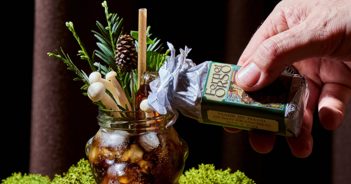 A hand sprays Fernet Orso from a small bottle into a bear-shaped glass filled with iced dark liquid and garnished with pine, mushrooms, and a pine cone.