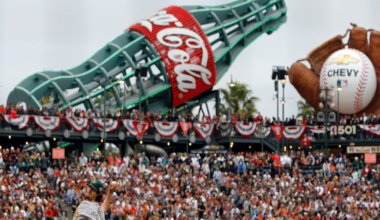 A baseball pitcher in mid-throw while a fielder readies as a large Coca-Cola bottle and baseball glove with ball dominate the stadium background.