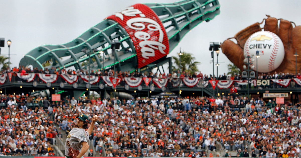 A baseball pitcher in mid-throw while a fielder readies as a large Coca-Cola bottle and baseball glove with ball dominate the stadium background.