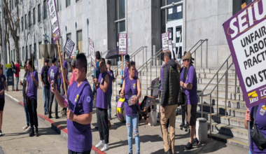 A group of people wearing purple shirts stand on a sidewalk outside a building, holding signs that say "SEIU Unfair Labor Practice On Strike."