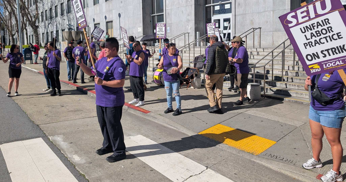 A group of people wearing purple shirts stand on a sidewalk outside a building, holding signs that say "SEIU Unfair Labor Practice On Strike."