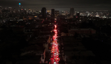 A long line of cars with bright red brake lights stretches down a dark city street at night, with illuminated buildings in the distant background.