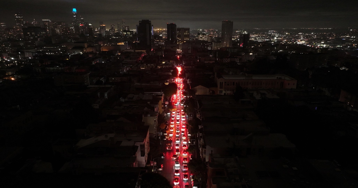 A long line of cars with bright red brake lights stretches down a dark city street at night, with illuminated buildings in the distant background.