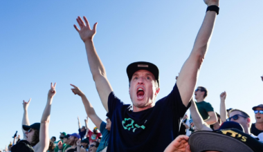 The image shows a baseball field lit up for a night game, surrounded by a cityscape, with a sunset over a water body and distant bridge and city skyline.
