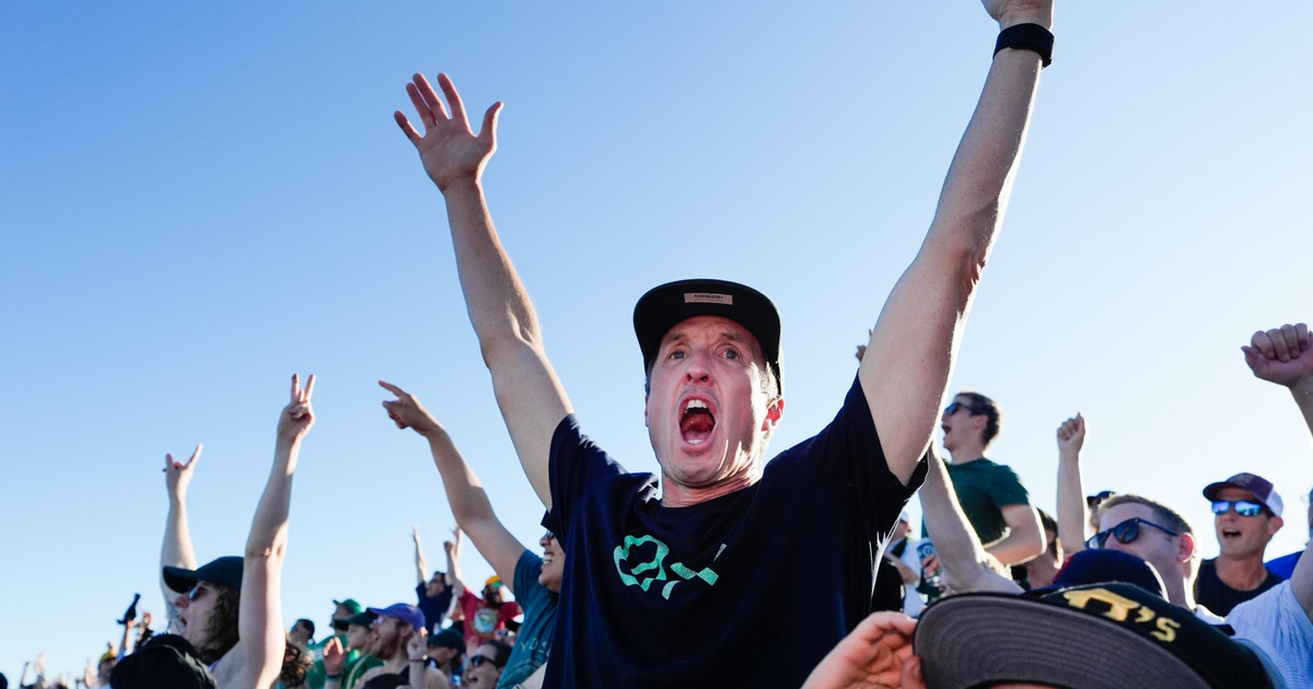 The image shows a baseball field lit up for a night game, surrounded by a cityscape, with a sunset over a water body and distant bridge and city skyline.