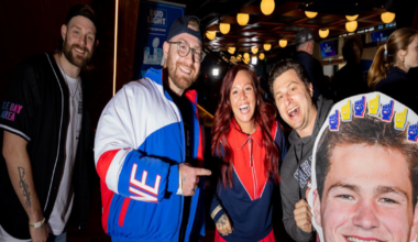Four people smile and pose indoors, with one holding a large cutout of a smiling man's face decorated with foam fingers on top.