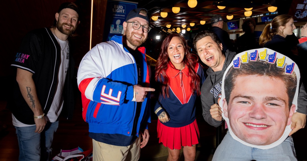 Four people smile and pose indoors, with one holding a large cutout of a smiling man's face decorated with foam fingers on top.