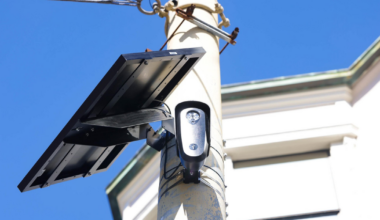 A solar panel and a small camera are mounted on a tall pole against a clear blue sky with part of a building in the background.