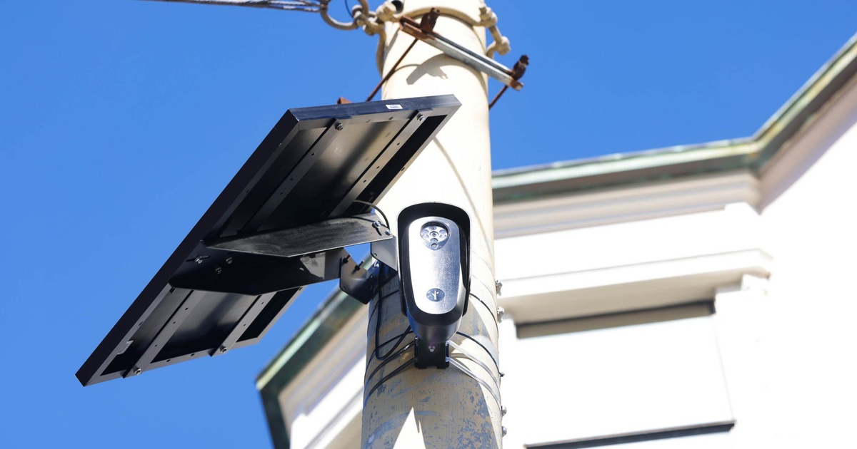A solar panel and a small camera are mounted on a tall pole against a clear blue sky with part of a building in the background.