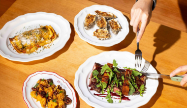 Four plates of food including grilled steak with greens, baked oysters, a stuffed pastry, and a side dish are arranged on a wooden table with a person cutting the steak.