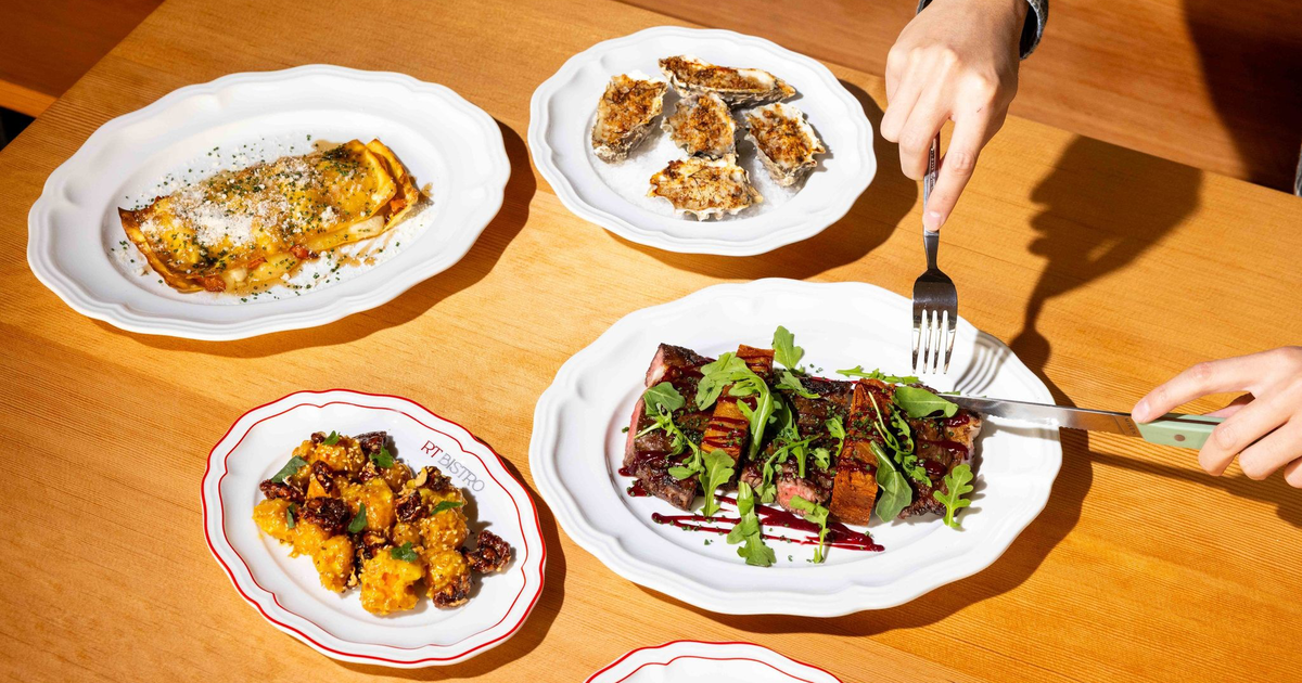 Four plates of food including grilled steak with greens, baked oysters, a stuffed pastry, and a side dish are arranged on a wooden table with a person cutting the steak.
