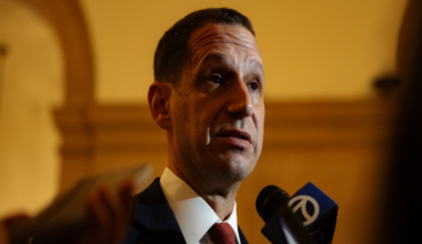 A man in a suit and red tie speaks into microphones, with a golden arch and warm lighting in the background.