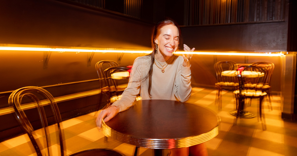 A young woman stands mid-street wearing a beige long-sleeve top, a black skirt, and black knee-high boots, with parked cars lining both sides.