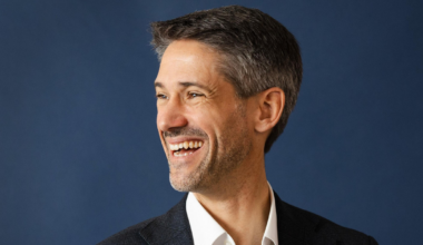 A man with short gray hair and a beard is smiling broadly, looking to his left, wearing a white shirt and a dark blazer against a blue background.